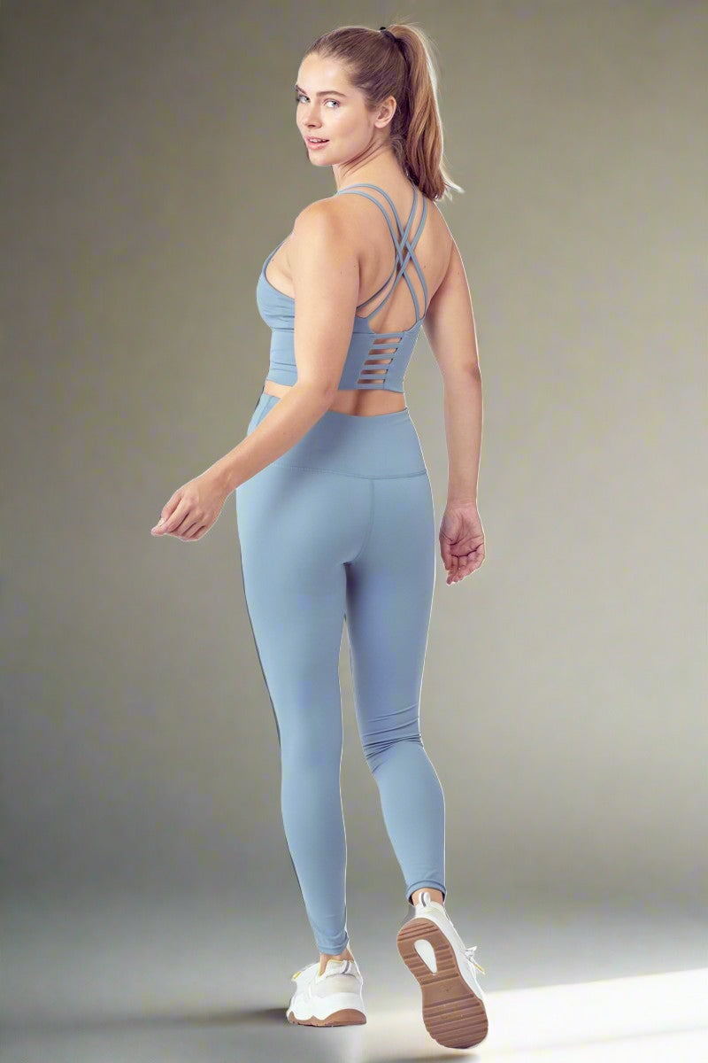 A person with long hair is wearing an Essential Activewear Set light blue sports bra with crisscross straps and matching high-waisted leggings, paired with white and yellow sneakers. They are standing and slightly turning back, smiling, against a plain white background.