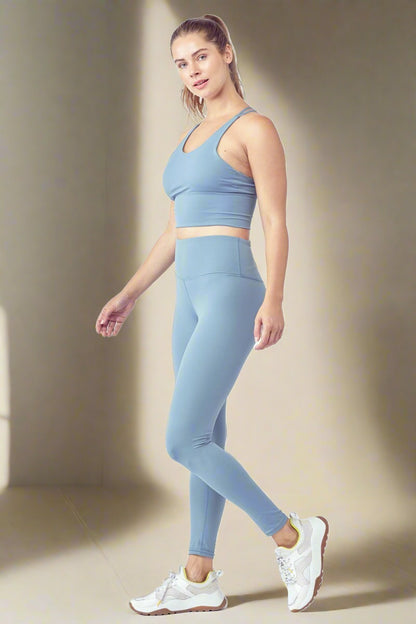 A woman with a ponytail poses against a plain background wearing the Essential Activewear Set, including a matching light blue cropped tank top and high-waisted leggings, along with white sneakers. She stands in a semi-profile position, looking at the camera with a relaxed expression.