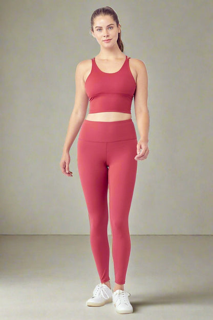 A woman stands against a white background wearing the Essential Activewear Set, consisting of a cropped tank top and high-waisted leggings. She has long hair pulled back in a ponytail and is wearing white sneakers. She appears relaxed and confident.