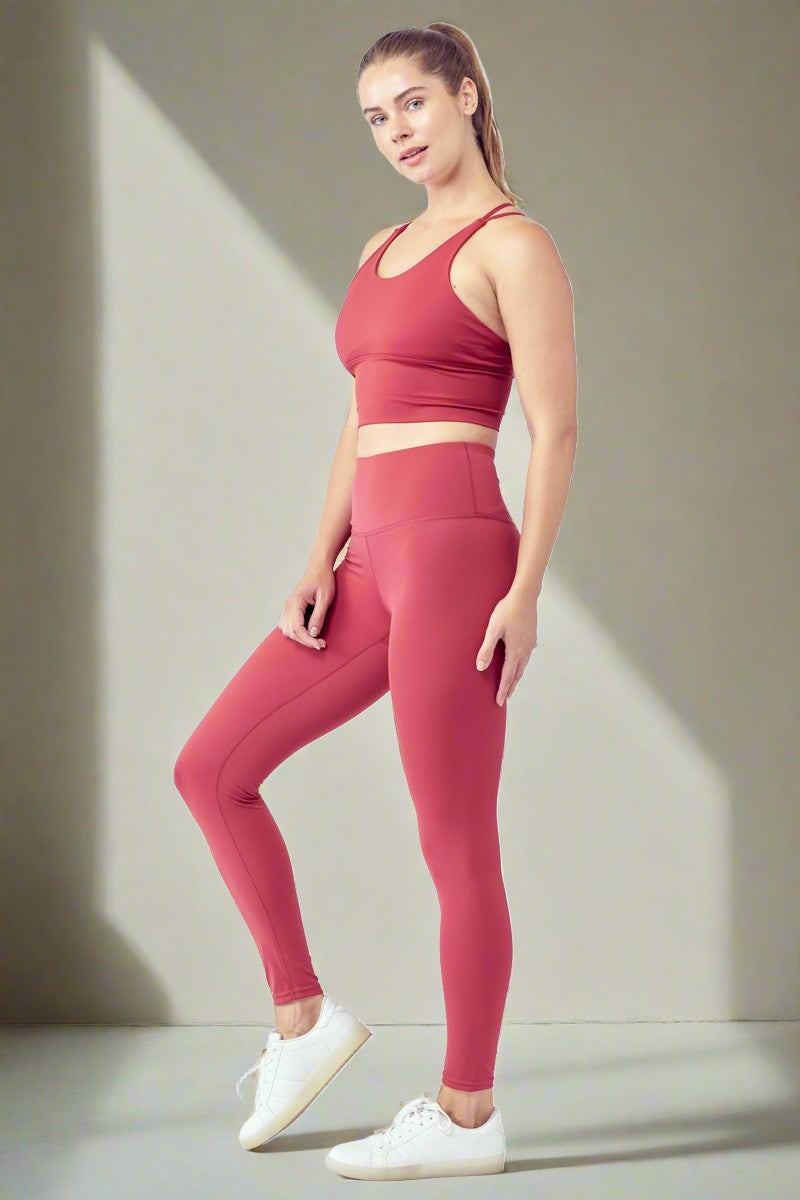 A woman stands facing slightly to the right against a plain white background. She is wearing a matching Essential Activewear Set, featuring a red sports bra and high-waisted leggings, along with white sneakers. Her hair is tied back and she has a neutral expression.