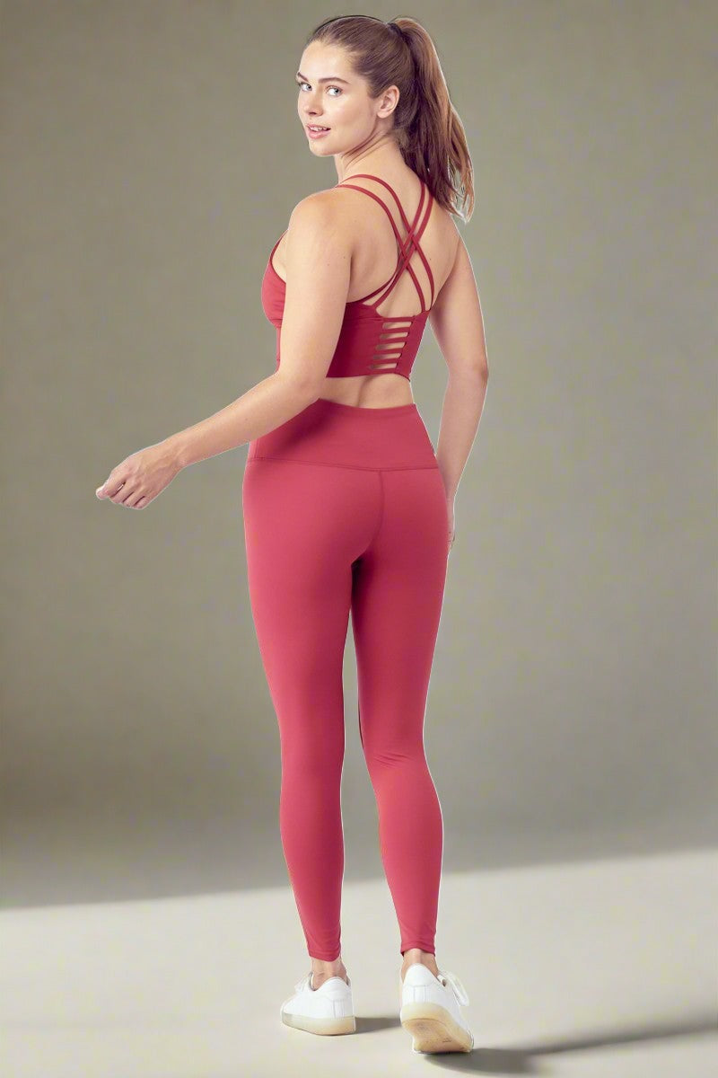 A woman stands facing away, looking over her shoulder. She is wearing a matching red Essential Activewear Set consisting of a strappy sports bra and high-waisted leggings, paired with white sneakers. Her hair is tied back in a ponytail. The background is plain white.