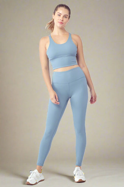 A woman dressed in a matching light blue cropped tank top and high-waisted leggings from the Essential Activewear Set stands confidently with her left hand resting on her thigh. She is wearing white sneakers with yellow accents and has her hair tied back in a ponytail, set against a plain, light background.