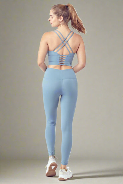 A woman with a ponytail is wearing the Essential Activewear Set, including a light blue strappy sports bra and matching high-waisted leggings, seen from the back. She is also sporting white sneakers with brown soles. The background is plain and light-colored.