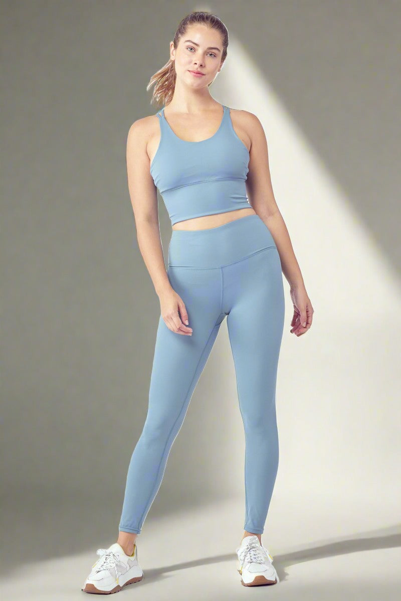 A woman is standing in a confident pose, wearing a light blue cropped tank top and matching high-waisted leggings from the Essential Activewear Set. She has white athletic shoes on her feet. The background is plain white, emphasizing her sporty ensemble.