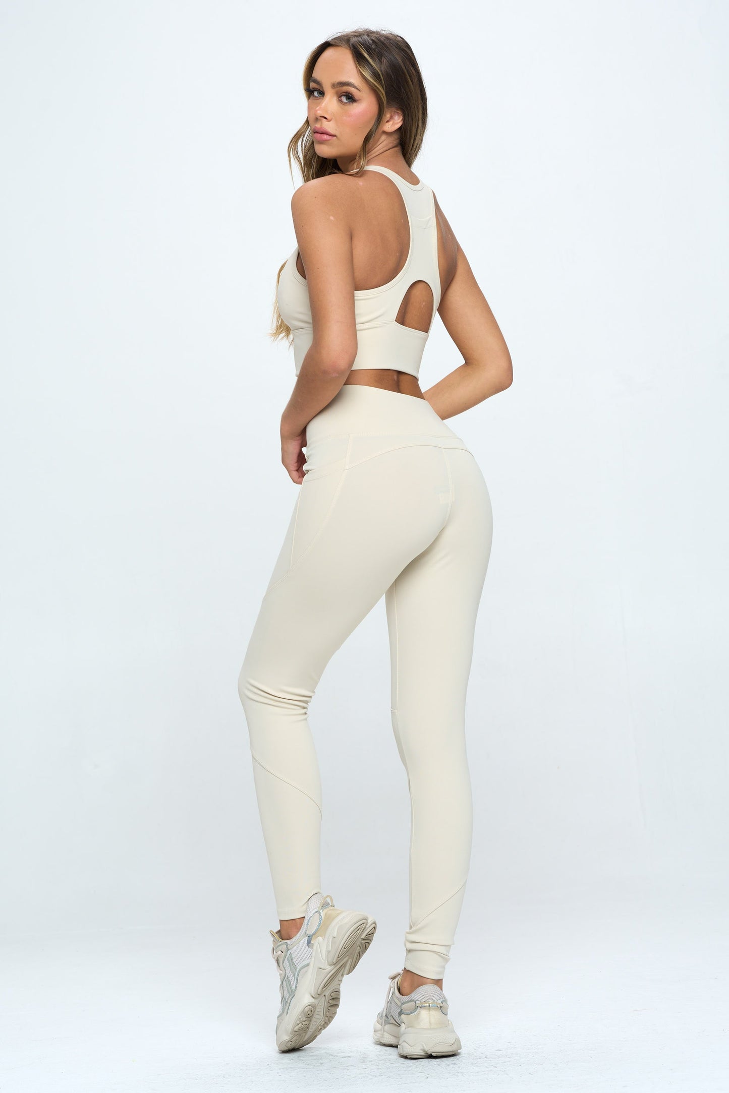 A person stands facing away from the camera, showcasing the Harmony Activewear Set in a beige sports bra and matching high-waisted leggings made from high-stretch fabric. The outfit is complemented by white sneakers. The plain white background emphasizes the athletic apparel.