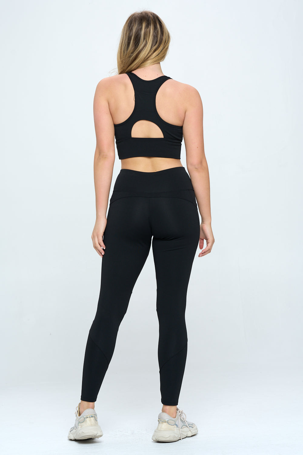 A woman with long, blonde hair stands facing away, wearing a black Harmony Activewear Set—a sports bra with a cutout in the back and high-waist, matching black leggings made from high-stretch fabric. She is also wearing white sneakers. The background is a plain white.