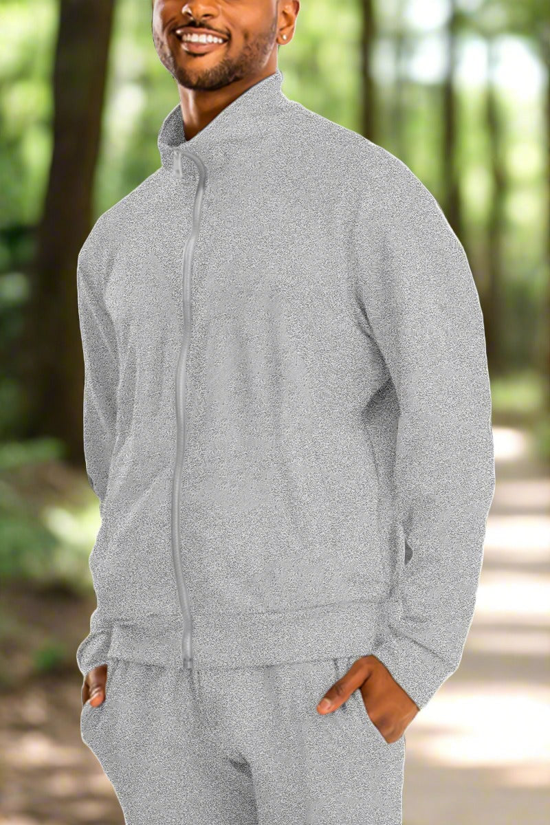 A man is smiling and wearing a light grey zip-up tracksuit with his hands in the pockets. The Essential Solid Track Jacket, with matching pants, features a high-collared jacket. Perfect for any casual wardrobe, this versatile style stands out against the plain white background.